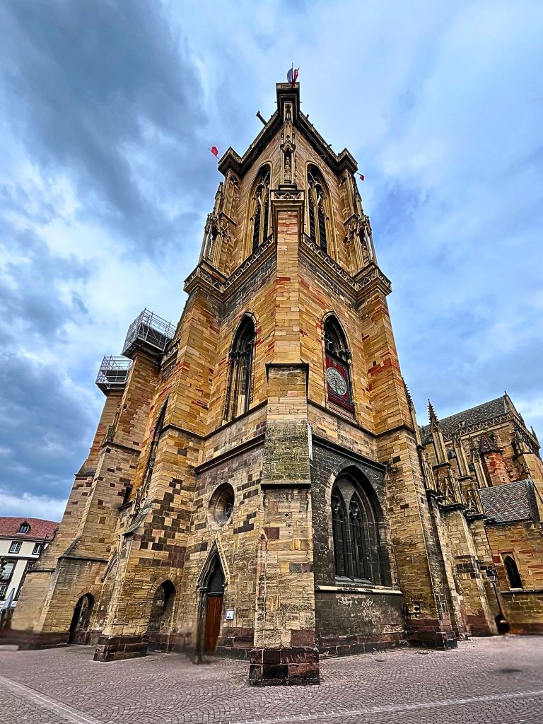 Close-up of the Gothic bell tower of the Collégiale Saint-Martin in Colmar, with detailed stone pinnacles, weathered sandstone, and two small flags fluttering near the top under a cloudy sky.