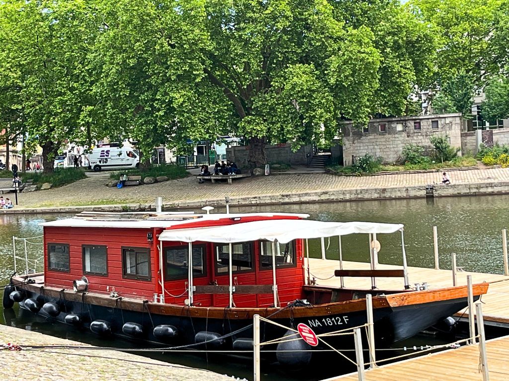 Wooden boat with weathered red walls moored along the Erdre River in Nantes, partially shaded by trees.