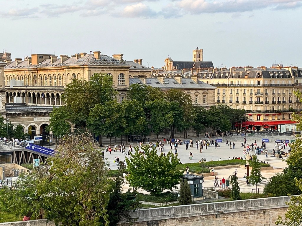 A wide view of Place Jean-Paul II in Paris, showing tree-lined walkways, Hôtel-Dieu’s cream-colored façade, and classic Parisian rooftops, with a red café awning on the far right.