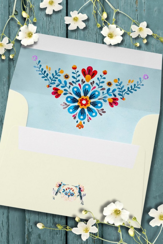 Cream wedding envelope featuring colorful Otomi-inspired floral liner in blue, red, and yellow, with small watercolor hummingbirds — part of the Mexican Folk Art Hummingbird Wedding Suite.