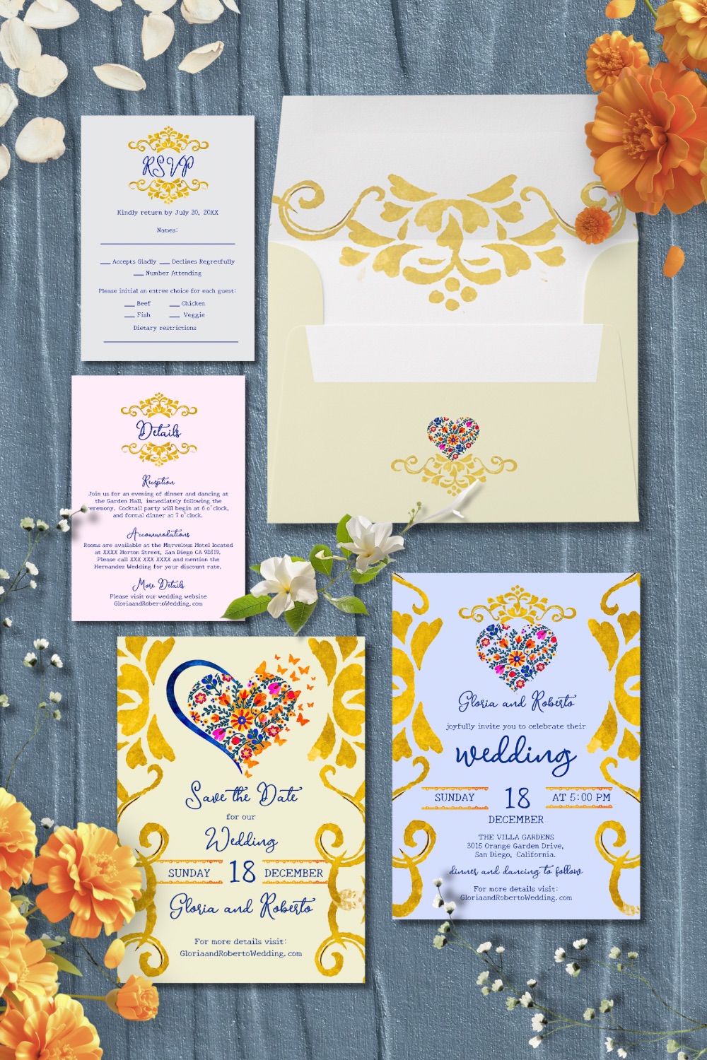 Lay flat view of a baroque floral heart Mexican wedding invitation suite with gold scrollwork, colorful folk heart motifs, RSVP, details card, and envelope on textured blue fabric